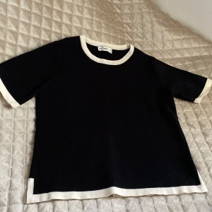 Svart stickad topp från Zara - Svart stickad topp från Zara med kontrasterande vita kanter vid halsringning, ärmslut och nederkant. Modellen har korta ärmar och en rak passform. Perfekt för dig som gillar en clean och stilren look