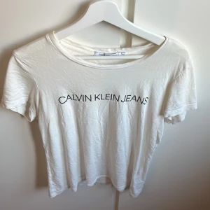 Vit t-shirt från Calvin Klein  - Vit t-shirt från Calvin Klein, med svart logotyptryck framtill. Klassisk rund halsringning och korta ärmar. Skön och enkel modell i mjuk bomull, Snygg basic som funkar till allt.