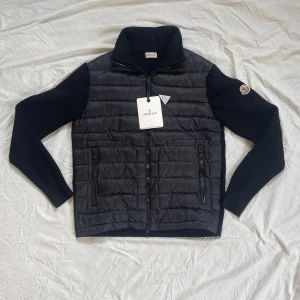 Moncler cardigan utav huva - Snygg svart pufferjacka från Moncler med quiltad front i polyester och stickade ärmar och rygg. Jackan har hög krage, dragkedja framtill och två fickor med dragkedja. Klassisk Moncler-logga på ärmen. Perfekt för dig som vill ha en stilren och sportig look. Den är helt ny och aldrig använd och är i mycket bra skick och suveränt kvalité. Priser kan även diskuteras!!