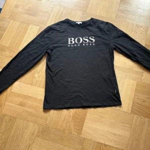 Svart långärmad tröja Hugo Boss - Svart långärmad tröja från Hugo Boss med stort vitt BOSS-tryck på bröstet. Klassisk rund halsringning och rak passform. Tillverkad i mjuk bomull som känns skön mot huden. Perfekt för dig som gillar stilrena och enkla plagg med tydlig logga.