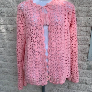 Rosa virkad kofta med knytband - Superfin rosa virkad kofta med långa ärmar och snyggt knytband med tofsar vid halsen. Koftan har ett öppet, luftigt mönster och vågig kant längs öppningen och nederkanten. Perfekt för lager-på-lager och ger en söt, bohemisk vibe.