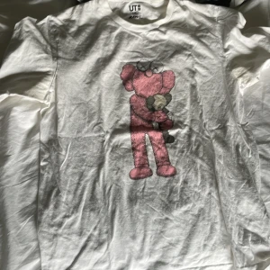 Vit KAWS x Uniqlo t-shirt XL - Vit t-shirt från Uniqlo i samarbete med KAWS, med en stor rosa KAWS-figur tryckt på framsidan. Klassisk passform och rund hals. Perfekt för dig som gillar streetwear och coola prints.
