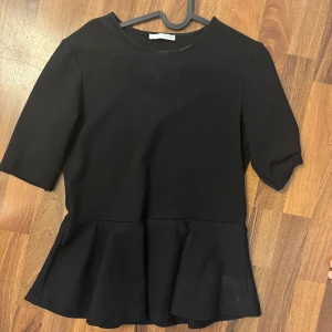 Svart peplumtop med halvlång ärm - En stilren svart topp med peplumvolang nedtill och halvlånga ärmar. Toppen har rund halsringning och är sydd i ett mjukt, lite stretchigt material som ger en snygg passform. Perfekt för dig som gillar enkel men trendig stil.