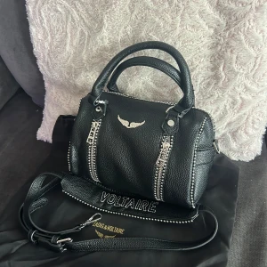 Svart handväska Zadig & Voltaire sunny - Cool svart handväska i skinn från Zadig & Voltaire med silverfärgade dragkedjor och nitar. Väskan har både handtag och en bred axelrem med texten VOLTAIRE. Snygg detalj med loggan i silver framtill och strukturerad yta.