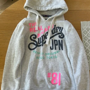 Grå Superdry hoodie med tryck - Snygg ljusgrå hoodie från Superdry med stort tryck i svart, rosa och turkos. De är knappt använd och verkligen skit snygg 😻😻men tyvärr lite liten för mig