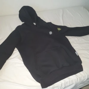 Svart hoodie från Stone Island - Svart hoodie från Stone Island med klassisk logga på ärmen och bröstet. Tröjan har huva, känguruficka och är tillverkad i mjuk bomull. Perfekt för dig som gillar streetwear och vill ha en stilren look.
