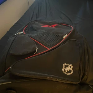Svart NHL hockey-resväska med hjul - Stor svart hockey-resväska med NHL-logga, röda detaljer och flera dragkedjefack. Väskan har utdragbart handtag och hjul för enkel transport. Perfekt för att få med sig all hockeyutrustning till och från träningen. Det medföljs även lite hockey puckar