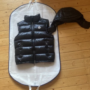 Svart Moncler dunväst med huva - Snygg svart dunväst från Moncler med glansig finish och avtagbar huva. Västen har två dragkedjefickor framtill och klassisk Moncler-logga på bröstet. Perfekt för lager-på-lager och håller dig varm med sin fluffiga dunfyllning. Stängs med dragkedja hela vägen. 
