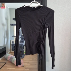 Svart ribbad topp från NA-KD - Svart långärmad topp från NA-KD med ribbad struktur och snörning på ena sidan. Snygg, figurnära passform och rund halsringning.