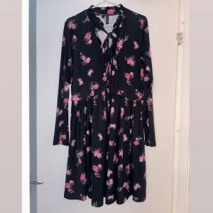 Svart blommig klänning från Rainbow - Supersöt svart klänning från Rainbow med rosa blommigt mönster, långa ärmar och knytning vid halsen. Klänningen har en lätt rynkad kjol och volangdetalj vid halsringningen. Perfekt för dig som gillar romantisk stil! Strl 40/42