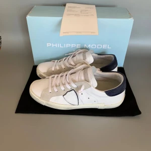 Vita sneakers från Philippe Model - Snygga vita sneakers från Philippe Model med detaljer i beige mocka och mörkblå häl. Skorna har klassisk snörning och en platt sula. Perfekta för dig som gillar stilrena och trendiga sneakers med exklusiv känsla.
