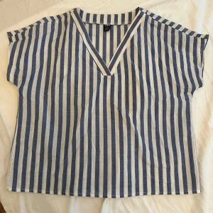 Randig blus med v-ringning från SHEIN - Säljer en luftig blus från SHEIN i storlek M. Toppen har blå och vita vertikala ränder, v-ringad hals och korta ärmar. Perfekt för en chill sommardag och enkel att matcha med jeans eller shorts.