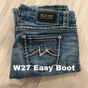 Miss Me Easy Boot jeans W27 - Snygga blå Miss Me Easy Boot jeans med coola kontrastsömmar och broderade bakfickor. Klassisk bootcut-modell med låg midja och slitningar för en avslappnad look. Jeansen är i mjukt denimtyg och har detaljerade nitar vid fickorna. Länd: 102 midja 38 benöppning 22 inseam 80