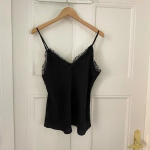Svart spetslinne från BikBok - Supersnygg svart linnetopp från BikBok med tunna axelband och v-ringning. Toppen har fina spetsdetaljer längs urringningen och är tillverkad i ett lätt, slätt material som ger en lyxig känsla. Perfekt att styla med jeans eller kjol för en trendig look.