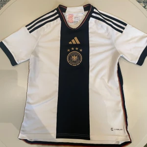 Tysklands fotbollströja Adidas 13-14 år/164 - Snygg Tyskland fotbollströja från Adidas i vitt med svart mittfält och guldiga detaljer. Tröjan har korta ärmar, klassiska Adidas-ränder på axlarna och broderat landslagsmärke framtill. Tillverkad i lätt och ventilerande polyester. Tröjan är självklart äkta, den är köpt ifrån adidas officiella hemsida. Enda defekten är på lägre höger sida (se bild 5).