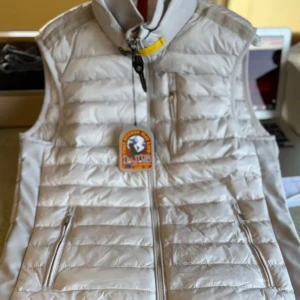 Vit dunväst från Parajumpers - Snygg och lätt dunväst från Parajumpers i en ljusvit nyans. Västen har quiltad design, hög krage med gul detalj och dragkedja framtill. Två fickor med dragkedja och en innerficka. Perfekt för lager-på-lager och streetstyle. Finns i fler storlekar 