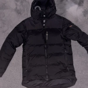 Svart dunjacka Sail Antarctic - Svart dunjacka från Sail Antarctic med huva och dragkedja framtill. Jackan har en quiltad design, logga på bröstet och ärmen samt praktiska fickor. Perfekt för kalla dagar när du vill hålla stilen och värmen.