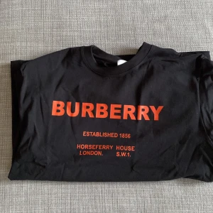 Svart Burberry t-shirt med röd logga - Svart t-shirt från Burberry i bomull med röd logotyp och text framtill. Klassisk rund halsringning och korta ärmar. Snygg och stilren design som sticker ut med den tydliga loggan. Perfekt för dig som gillar exklusiva streetwear-plagg. Storlek M, men som L