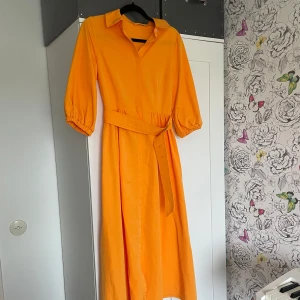 Orange långklänning  - Min älskade retroklänning, 60s känsla med knappar och ett brett bälte i midjan, snygg krage och 3/4 ärmar. Använd fåtala gånger, en extra tryckknapp insydd mitt på bysten.