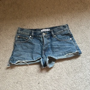 Low waist jeans shorts  - Snygga blå low waist  jeansshorts från & Other Stories med råa kanter och klassisk femficksdesign. Shortsen har normal passform, bälteshällor och knappstängning framtill. Perfekta för sommardagar och ger en avslappnad vibe.