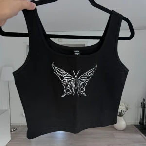 Svart croppad linnetopp med fjäril - Svart croppad linnetopp med breda axelband och en glittrig fjäril framtill i silvriga stenar. Toppen har en enkel, rak passform och är perfekt för dig som gillar statement-detaljer. Materialet är mjukt och stretchigt <3 Passar S/M! Önskar du veta exakta mått? Tveka inte att höra av dig!
