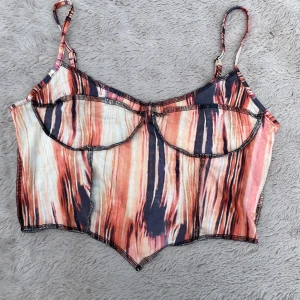 Färgstark korsett-topp från SHEIN - Säljer en snygg korsett-topp från SHEIN i storlek M. Toppen har tunna axelband och ett coolt randigt mönster i orange, beige, svart och vitt. Den har markerade sömmar och en spetsig nederkant som ger en edgy vibe. Perfekt för dig som vill sticka ut!