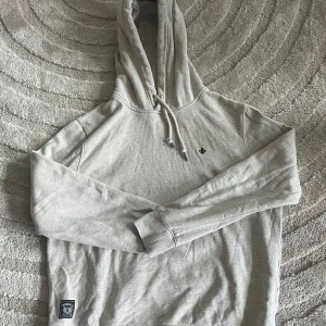 Ljusgrå hoodie från Morris - Snygg ljusgrå hoodie från Morris med broderad logga på bröstet och patch nertill. Klassisk modell med huva och snörning, samt långa ärmar och ribbade muddar. Perfekt för chill dagar eller när du vill ha en clean look.