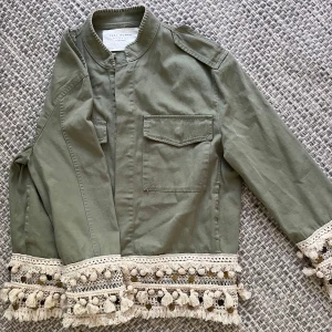 Grön overshirt med boho-detaljer Zara - Snygg militärgrön overshirt från Zara med unika virkade detaljer och tofsar i beige längs ärmslut och nederkant. Jackan har rak passform, bröstficka och ståkrage. Perfekt för dig som gillar boho-vibes och vill sticka ut lite extra.
