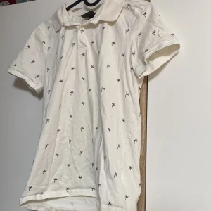 Vit pikétröja med palmmönster H&M XS - Vit pikétröja från H&M i slim fit med små svarta palmmönster över hela tröjan. Klassisk krage och korta ärmar, perfekt för sommaren. Tillverkad i 100% bomull för en skön och luftig känsla.