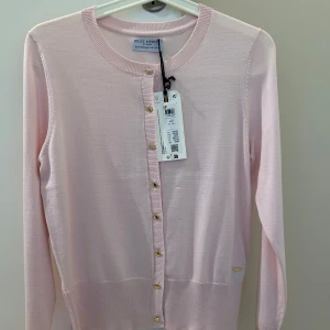  Ny Ljusrosa kofta från Holly & Whyte - Supersöt ljusrosa kofta från Holly & Whyte med guldiga knappar framtill. Den är tunnstickad i mjuk bomull, har rund hals och långa ärmar. Perfekt att slänga över en topp för en chill och fräsch look.