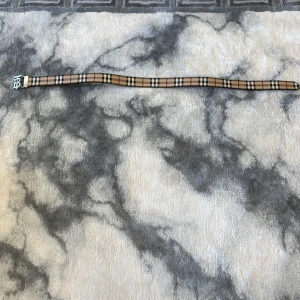 Burberry vändbart bälte med silverdetalj - Snyggt vändbart bälte från Burberry med klassiskt rutigt mönster på ena sidan i beige, svart och vitt samt helsvart på andra sidan. Bältet har en stor silvrig spänne med Burberrys ikoniska 'B'-logga. Perfekt accessoar för att lyfta din outfit.