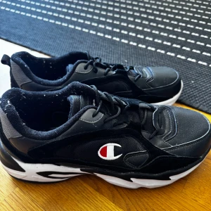 Svarta sneakers från Champion - Snygga svarta sneakers från Champion med vit och röd logga på sidan. Skorna har en chunky vit sula, svarta och grå detaljer samt snörning. Ovandelen är i en mix av syntet och mesh för en sportig vibe. Perfekta för dig som gillar streetstyle och bekväma skor.