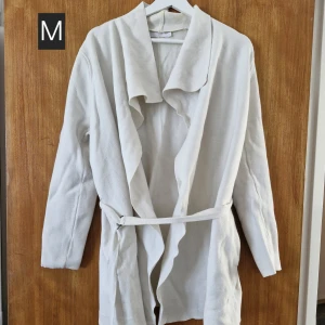 Vit kofta med bälte, M - Mysig vit kofta med långa ärmar och snygg omlottkrage. Jackan har ett bälte i midjan för en skön och avslappnad passform. Perfekt för lager-på-lager och chill dagar.