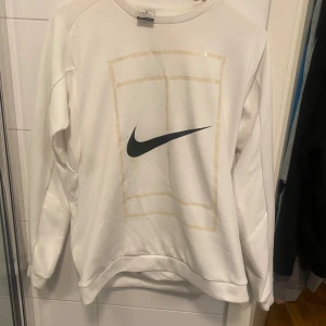 Vit Nike Dri-Fit sweatshirt M - Vit långärmad Nike Dri-Fit sweatshirt med svart swoosh-logga och grafiskt tryck framtill. Tröjan är tillverkad i ett lätt och ventilerande polyestermaterial, perfekt för sportiga dagar eller chill outfits. Klassisk passform och rund hals.