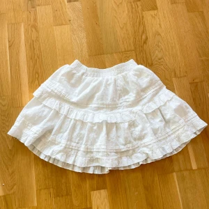 Vit volangkjol med resårmidja - Superfin vit kortkjol med flera volanger och resår i midjan. Kjolen har diskreta prickar i tyget och är fodrad för extra komfort. Perfekt för en somrig look och lätt att matcha med olika toppar.