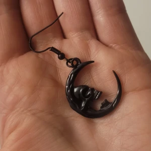  Helt nya Svarta örhängen med dödskalle och måne - Helt nya,svarta örhängen i form av en halvmåne med en detaljerad dödskalle i mitten. De har en blank finish och hängs på krok. Perfekta för dig som gillar alternativ stil och vill sticka ut med unika accessoarer.
