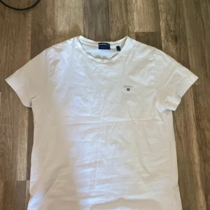 Vit t-shirt från GANT, storlek S - Vit t-shirt från GANT i klassisk modell med rund hals och korta ärmar. T-shirten har GANT-logga broderad på bröstet och är tillverkad i mjuk bomull. Perfekt basic-plagg med clean stil och regular fit.