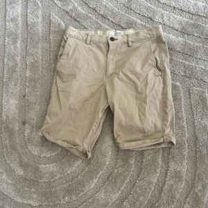 Beige shorts från Jack & Jones - Snygga beige shorts från Jack & Jones med klassisk passform och uppvikta benslut. De har fickor fram och bak, bälteshällor och stängs med knapp och dragkedja. Perfekta för varma dagar och enkla att matcha med det mesta.