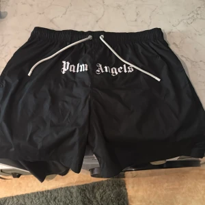 Svarta Palm Angels badshorts  - Svarta shorts från Palm Angels i storlek XL med vit logga framtill och vita snören i midjan. Tillverkade i 100% polyester. Elastisk midja för skön passform. 100% äkta kvitto kan visas köpt på farfetch.