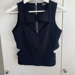 Svart croppad topp med cut-outs - Säljer en svart ärmlös topp med coola cut-outs och dragkedja både fram och bak. Toppen har en modern design med markerad midja och passar perfekt till en trendig outfit. Står storlek L men upplever den mer som M.