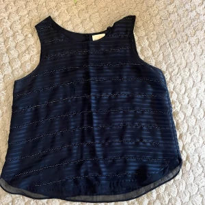 Mörkblå randig blus Holly & Whyte - Snygg mörkblå blus från Holly & Whyte by Lindex. Ärmlös modell med tunna horisontella ränder och diskreta prickdetaljer. Lätt och luftig, perfekt till jeans eller kjol. Stängs med knapp i nacken för en stilren look. Några kristaller är borta men inget man märker