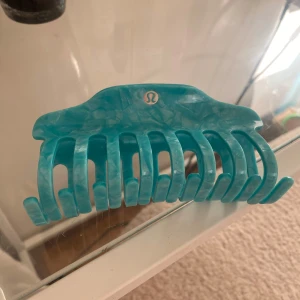 Turkos hårklämma från Lululemon - Snygg turkos hårklämma från Lululemon med marmorerad finish och klassisk logga framtill. Klämman har robust fjäder och breda taggar som håller håret på plats hela dagen. Perfekt accessoar för att snabbt fixa frisyren. Aldrig använd