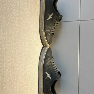 Svarta sneakers med fågelbrodyr - Svarta sneakers i skinn med grå snörning och vit sula. På sidan finns en broderad fågel i vitt som ger en unik touch. Klassisk låg modell som passar till det mesta och har en stilren vibe.