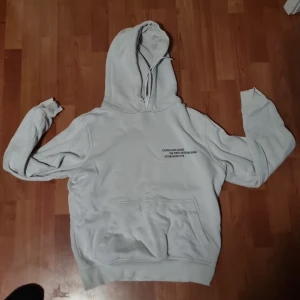 Vit hoodie från Calvin Klein Jeans - Snygg vit hoodie från Calvin Klein Jeans med tryck på bröstet och en stor grafisk print på ryggen. Tröjan har huva med snörning, känguruficka och ribbade muddar. Perfekt för dig som gillar streetstyle och vill ha något bekvämt men ändå stilrent.