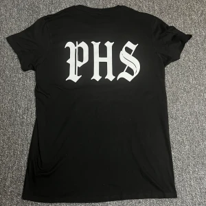 Svart PHS Support t-shirt - Svart t-shirt med stort vitt PHS-tryck på ryggen och mindre tryck framtill med knogjärn, lagerkrans och texten 'SUPPORT'. Klassisk rund hals och korta ärmar. Skön passform och tillverkad i mjukt bomullstyg. Perfekt för dig som gillar streetwear och tydliga statement-plagg.