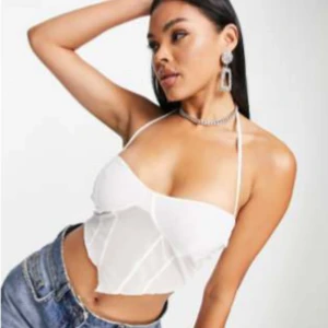 Vit topp  - Säljer en vit topp med halterneck och croppad passform. Toppen har tunna band som knyts i nacken och markerade sömmar framtill för en snygg siluett. Perfekt att styla med jeans eller kjol för en trendig look.