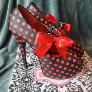 Bordello Teeze svarta med röda prickar - Unika pumps från Bordello i svart satin med röda prickar och stora röda rosetter framtill. Skorna har 14,6 cm hög klack, peep-toe och dold platåsula för extra attityd. Perfekta för dig som vill sticka ut med ett lekfullt och retroinspirerat uttryck. Använda endast en gång. Originalpris 893 kr