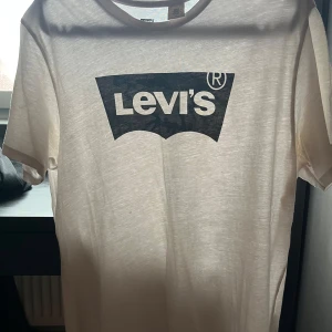 Vit Levi's t-shirt med logga - Vit t-shirt från Levi's i storlek Youth L med militär Levi's loggan tryckt på bröstet. T-shirten har rund halsringning och korta ärmar. Perfekt för en clean och avslappnad stil. Inte genomskinlig fönstret bakom får de o se ut som det.