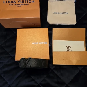Svart Louis Vuitton monogram bälte - Snyggt svart bälte från Louis Vuitton med klassiskt monogrammönster och stor LV-logga i matt svart metall som spänne. Bältet är gjort i präglat skinn och har en exklusiv vibe som verkligen sticker ut. Perfekt accessoar för att lyfta din outfit.