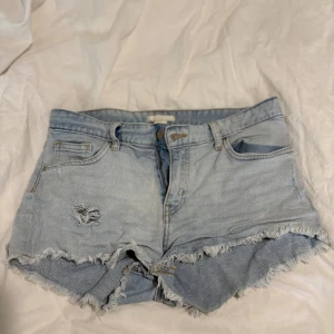 Ljusblå jeansshorts med fransig kant - Snygga ljusblå jeansshorts med fransig rå kant och slitningar framtill. Klassisk femficksmodell med bälteshällor och dragkedja. Perfekta för varma dagar och ger en avslappnad vibe.